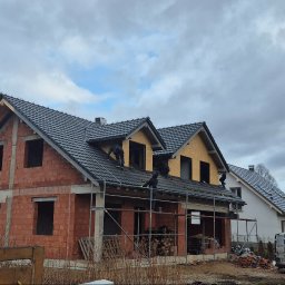 SYLWIA NAJDA - Budowa domu z czerwonej cegły w trakcie krycia dachu ciemną dachówką. Na dachu pracownicy. W tle drugi dom i zaparkowane samochody. Pochmurne niebo.