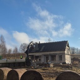 SYLWIA NAJDA - Budowa domu w trakcie: murowane ściany, kryty dachówką dach, dźwig podający materiały. W tle łąka z balotami siana i błękitne niebo z chmurami.