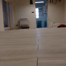 Układanie jasnych, podłużnych płytek podłogowych w nowym pomieszczeniu. Widoczne krzyżyki dystansowe, przygotowane kable w ścianach i klimatyzator.