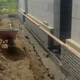 Wznoszenie ścian zewnętrznych z bloczków betonowych i cegły klinkierowej, widoczna izolacja termiczna i wykop, taczka w tle.