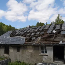 Częściowo zdjęty dach z łupka na starym domu z odsłoniętą drewnianą konstrukcją więźby dachowej, satelity na dachu, tło z drzewami i niebem z chmurami.