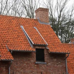 Czerwona dachówka ceramiczna na ceglanym domu z oknami dachowymi i kominem, widoczna z bliska na tle drzew bez liści.