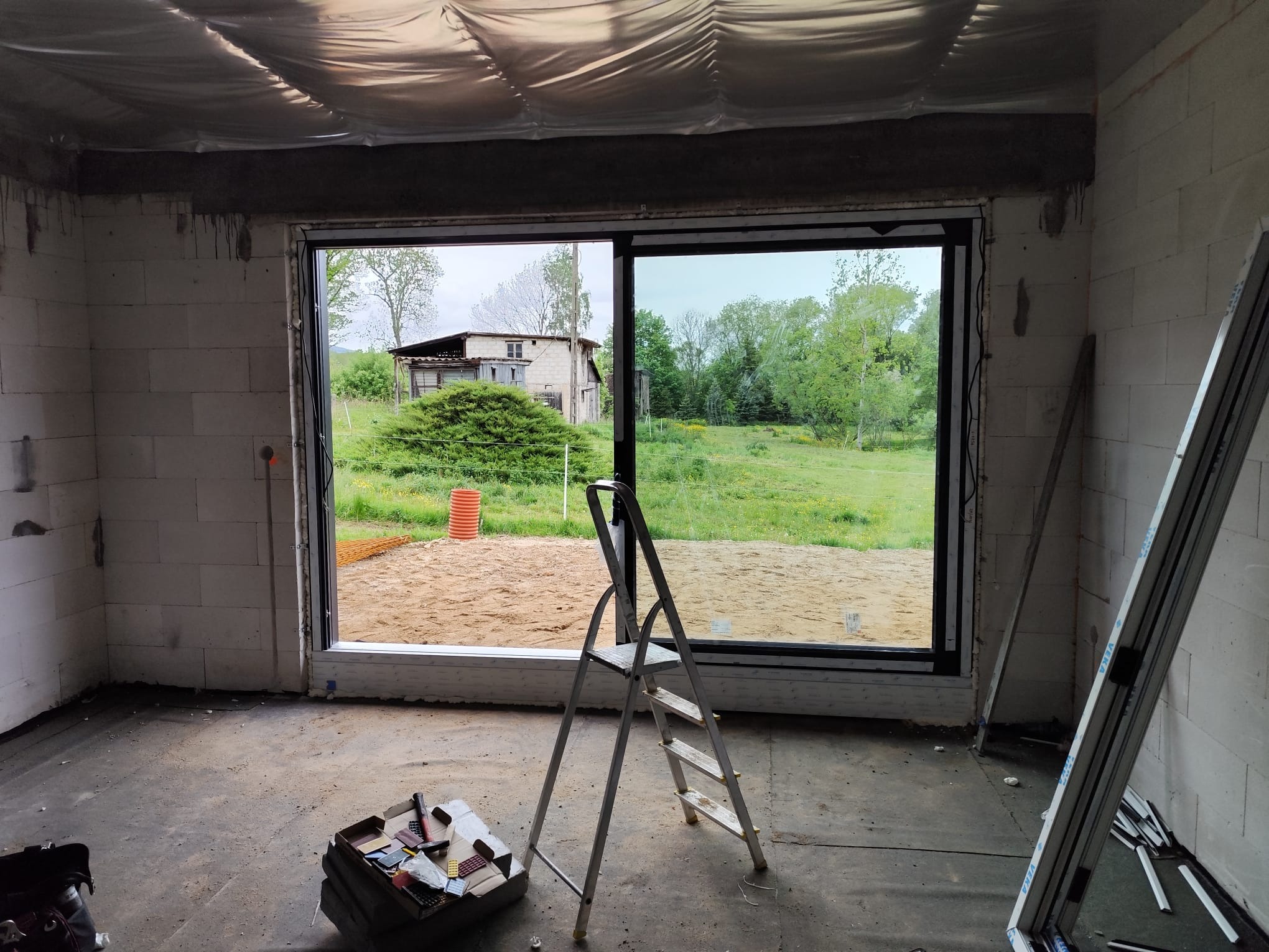 Wnętrze surowego domu z pustaków, z zamontowanym oknem z widokiem na wiejski krajobraz. W pomieszczeniu drabina i próbki materiałów wykończeniowych.