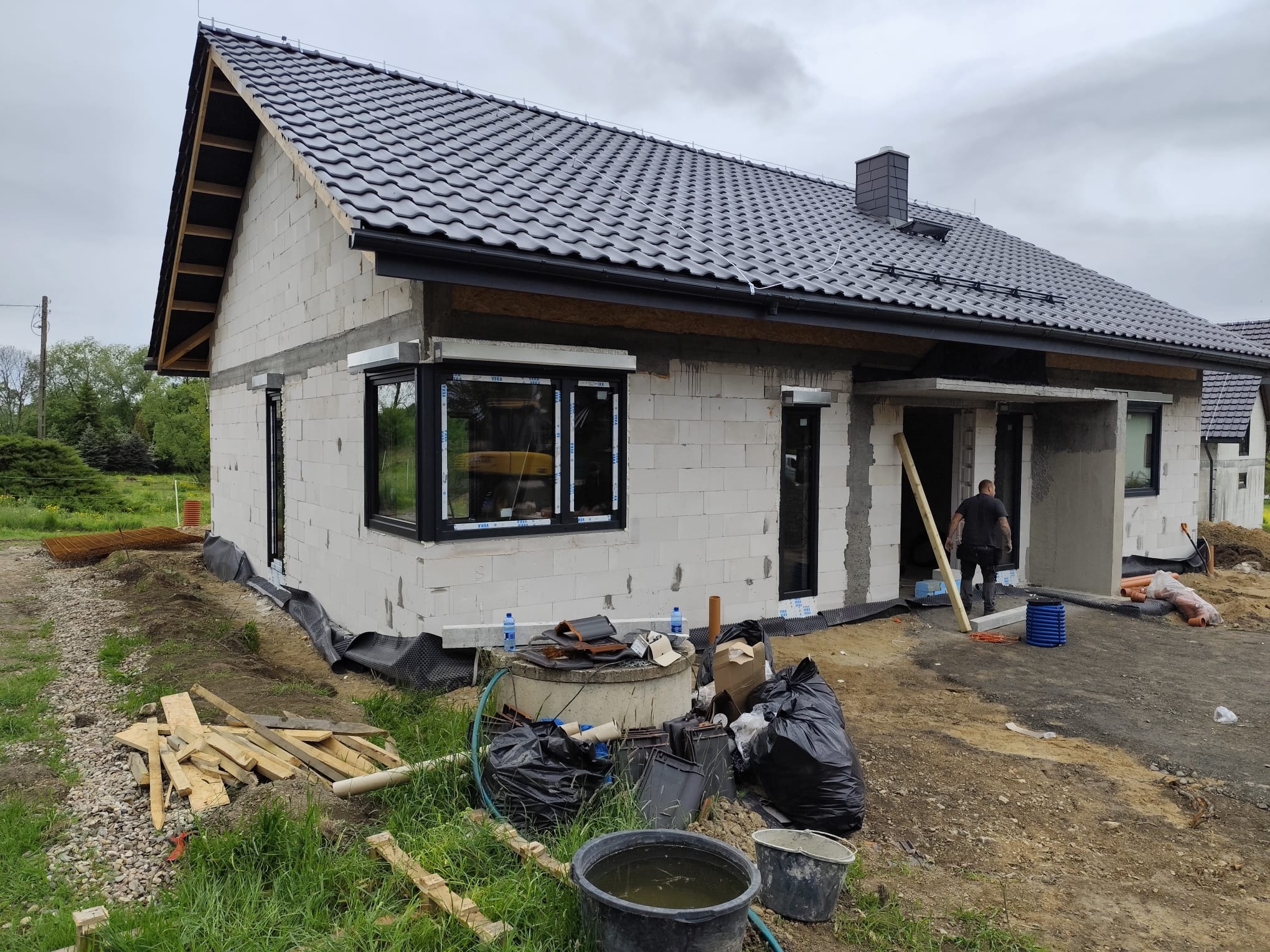 Budowa domu z białych bloczków, widoczny dach z ciemnej dachówki, czarne okna. Na pierwszym planie teren budowy z materiałami i narzędziami.