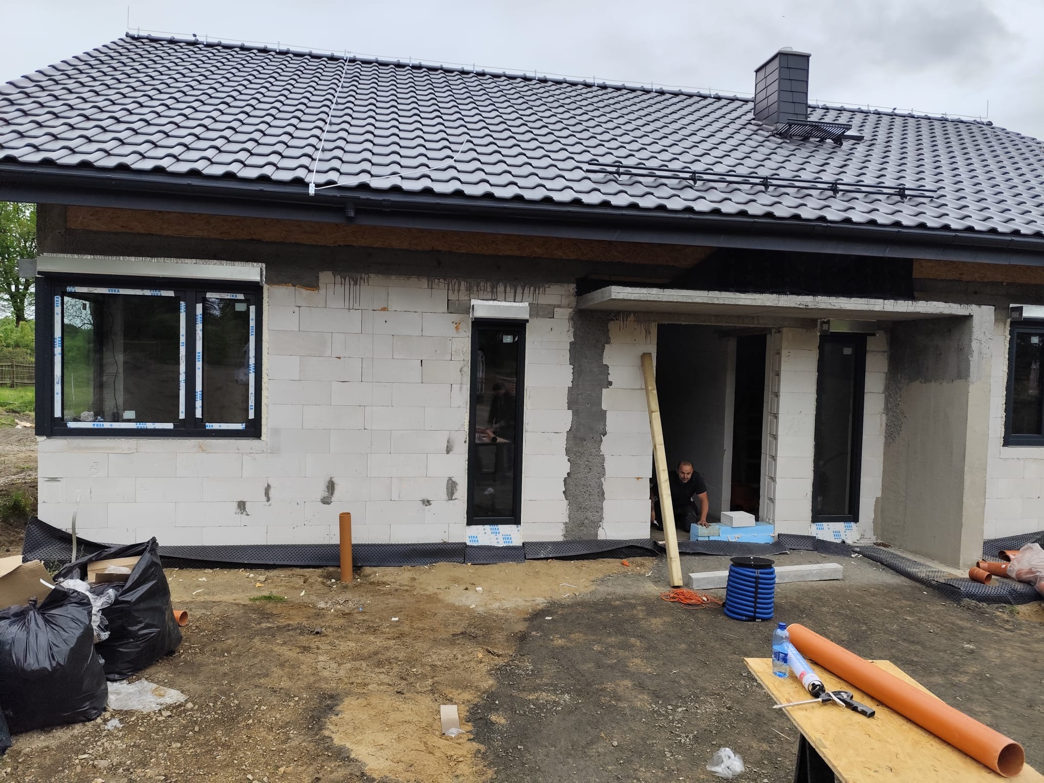Budowa domu w trakcie: ściany z białego bloczka, czarna dachówka, okna z czarnymi ramami. Na pierwszym planie rury i narzędzia, w tle pracownik.