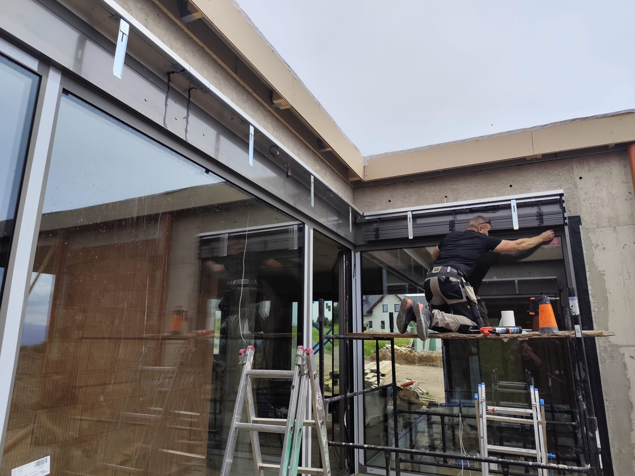 Montaż rolet zewnętrznych na surowym budynku z dużymi oknami. Pracownik na rusztowaniu, widoczne narzędzia i drabiny. Budynek w stanie deweloperskim.