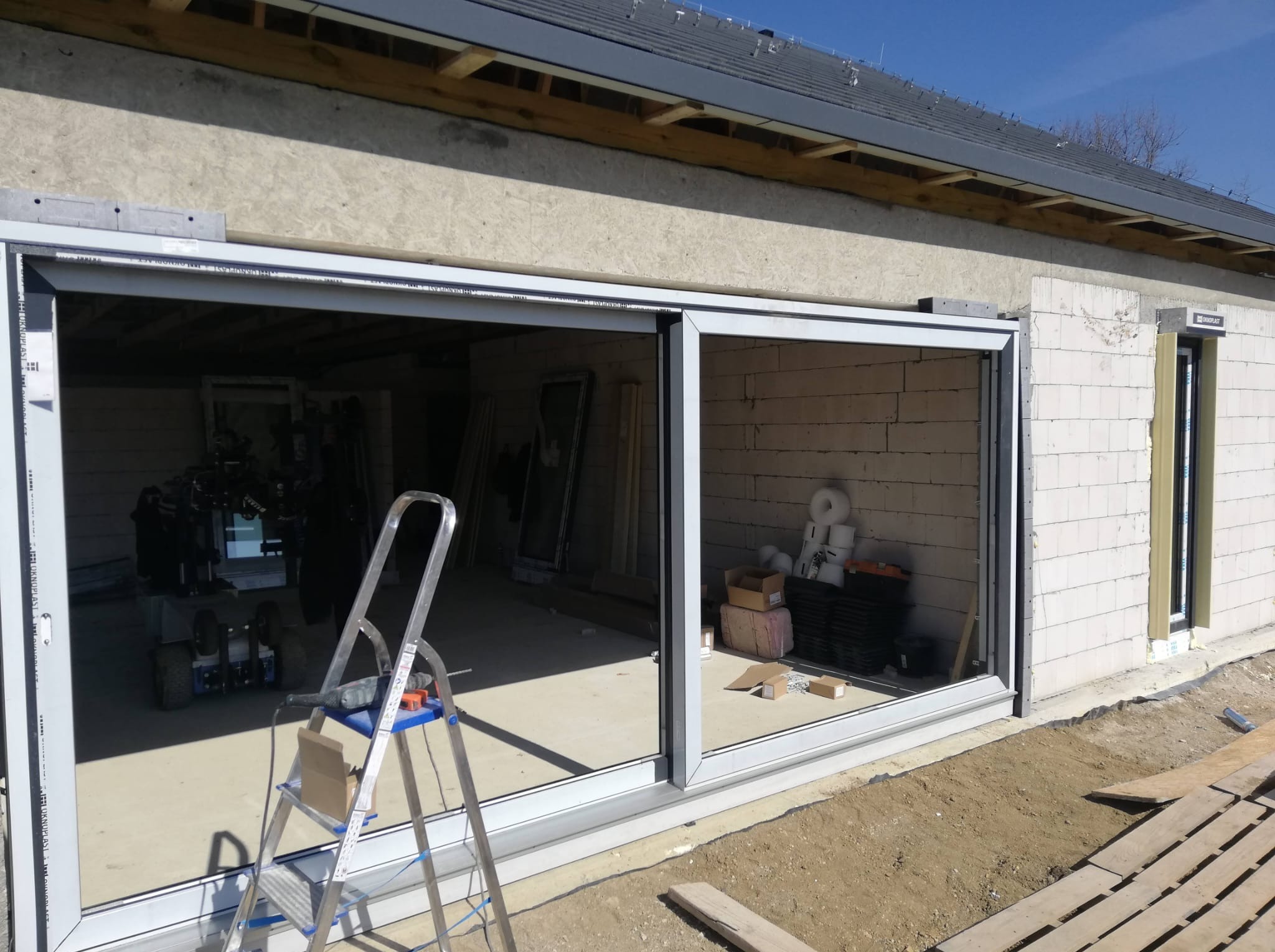 Montaż okna przesuwnego w surowym budynku z białego bloczka. Widoczna drabina, narzędzia i materiały budowlane. Zewnętrzna część budynku z drewnianymi elementami.