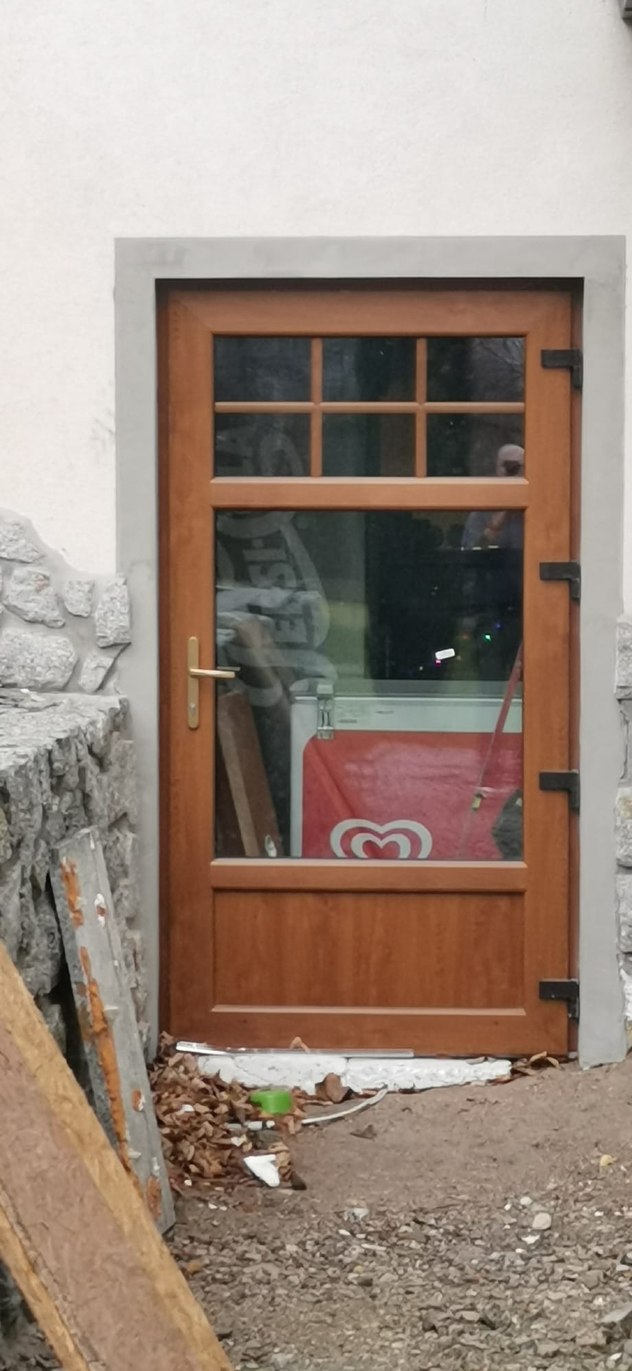 Brązowe drzwi wejściowe z szybą, widoczne z zewnątrz. W tle wnętrze z lodówką i elementami dekoracyjnymi. Częściowo widoczny kamienny mur i ziemia.