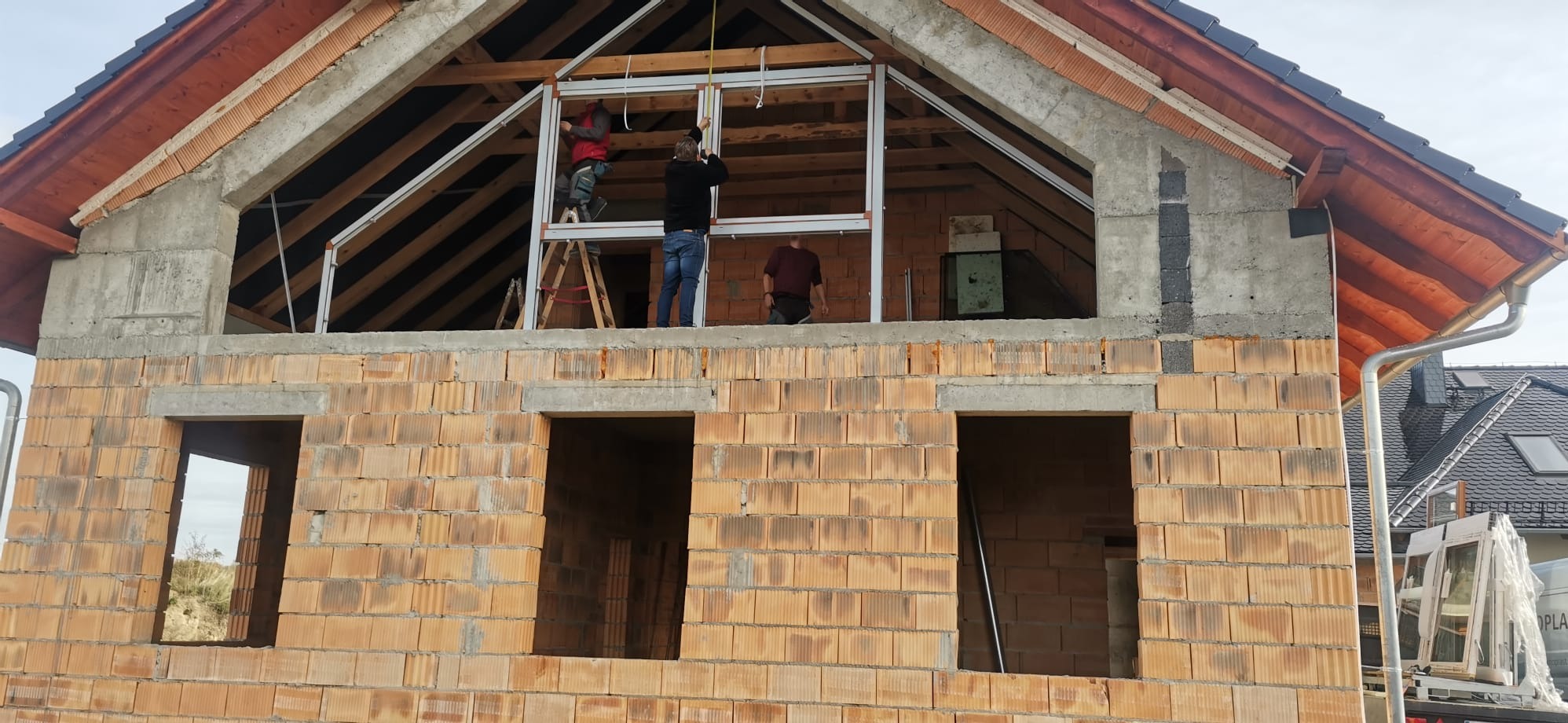 Budowa domu z czerwonej cegły: robotnicy montują okna na poddaszu. Widoczne drewniane elementy dachu i okna czekające na montaż. Detal: rynna po prawej.