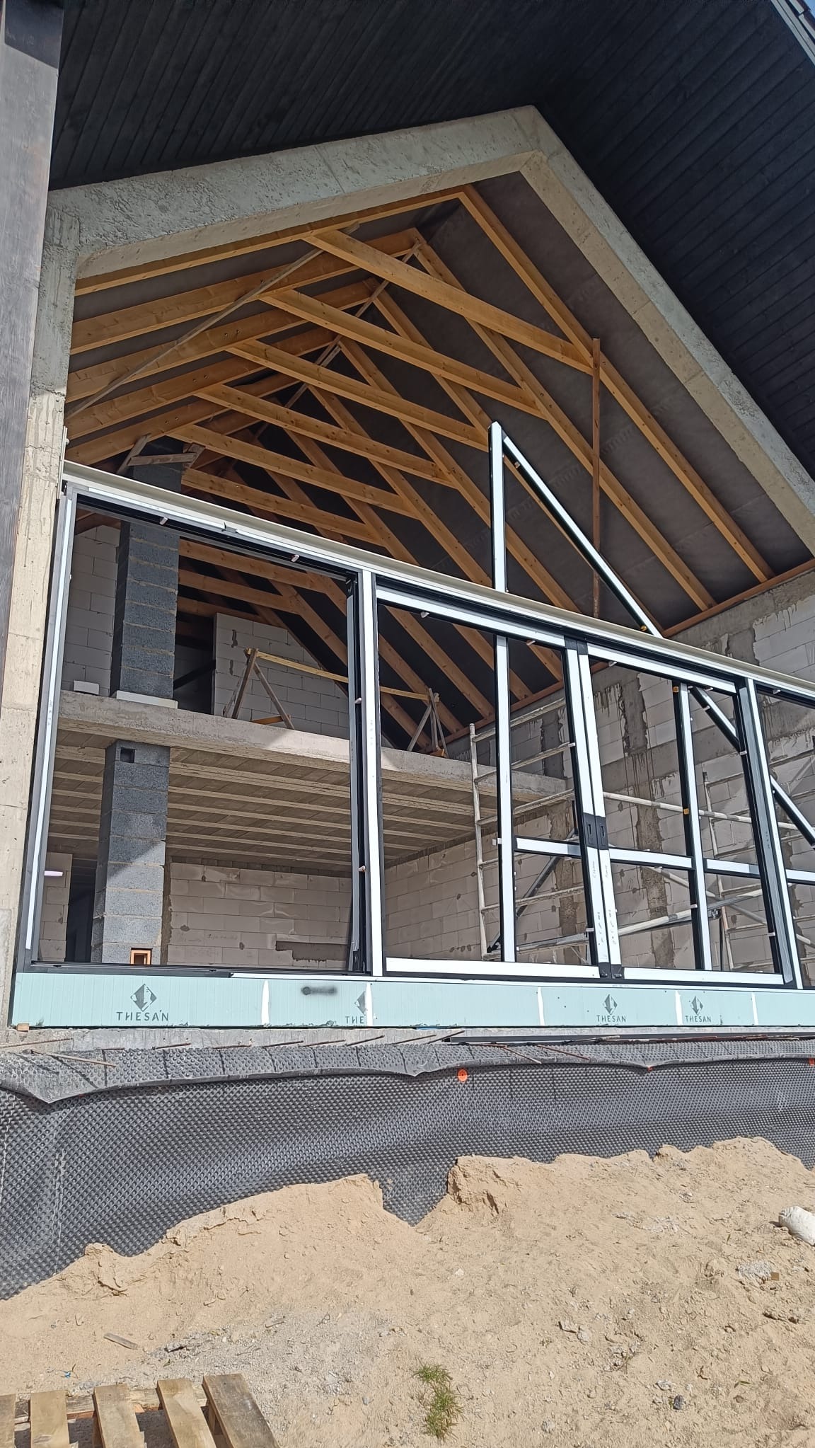 Surowy stan domu z widoczną konstrukcją dachu, oknami przesuwnymi i izolacją fundamentów. Widoczny szkielet budynku w trakcie budowy.