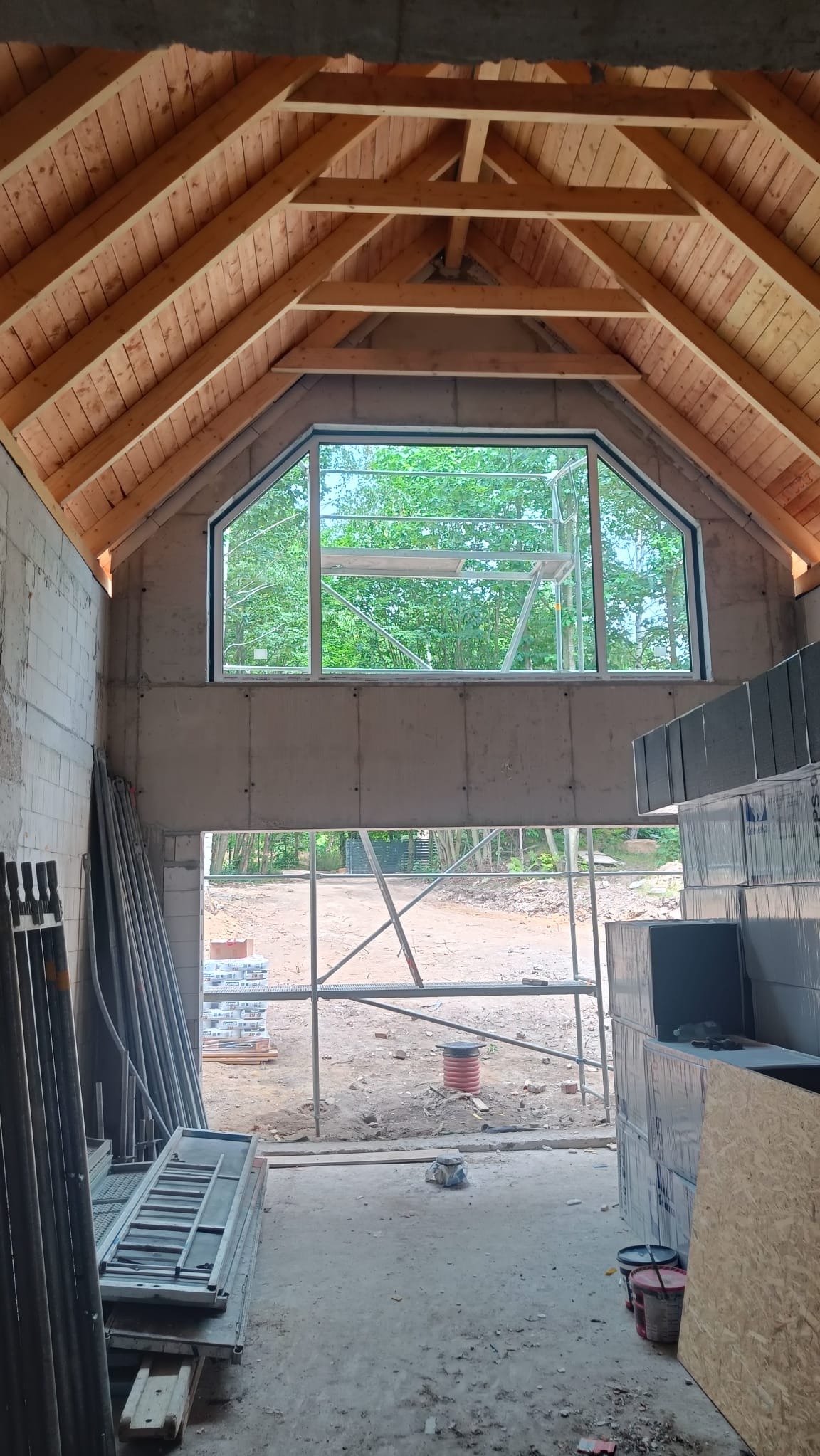 Surowy stan domu w budowie: drewniany dach z oknem w szczycie, widok na rusztowania i zieleń. Elementy konstrukcyjne i materiały budowlane wewnątrz.