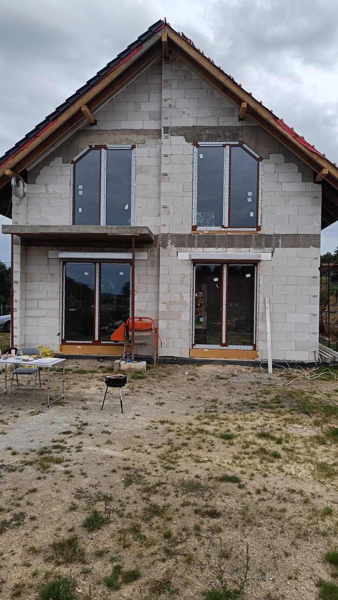 Surowy stan domu z białego bloczka, z zamontowanymi oknami i częściowo wykończonym dachem. Widoczny mikser budowlany i grill na pierwszym planie.