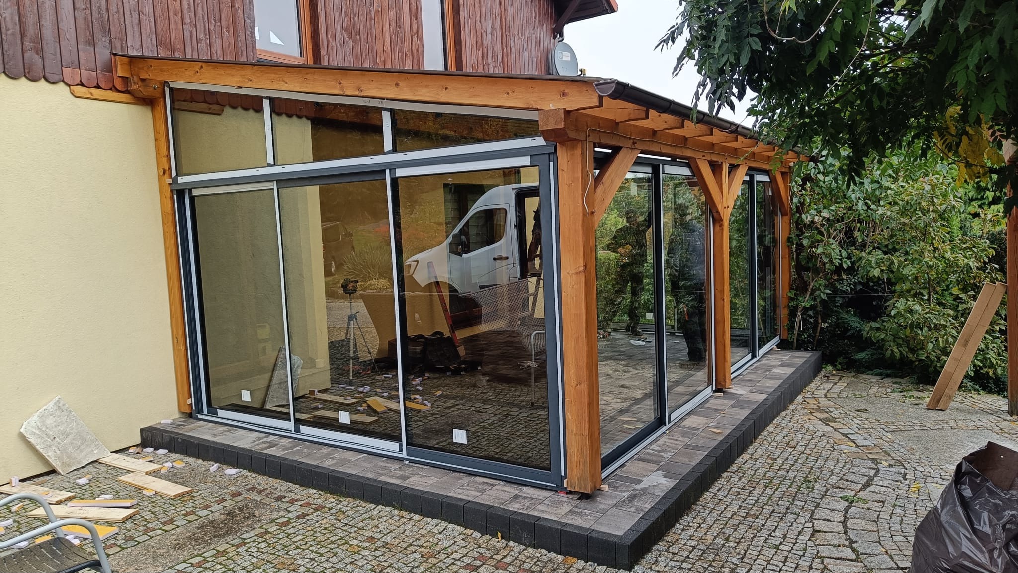 Zadaszony taras z przesuwnymi szklanymi ścianami, drewnianą konstrukcją i kamienną posadzką. Widoczny biały samochód dostawczy w odbiciu szyb.