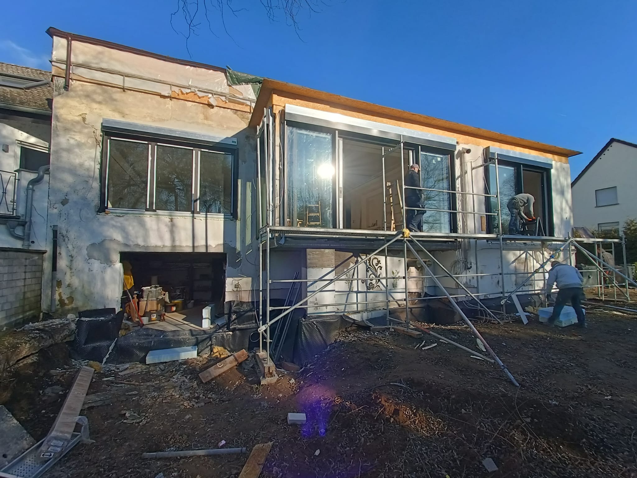 Remontowany dom jednorodzinny z rusztowaniem. Widoczne nowe okna, prace wykończeniowe elewacji i robotnicy. Słoneczny dzień, błękitne niebo.