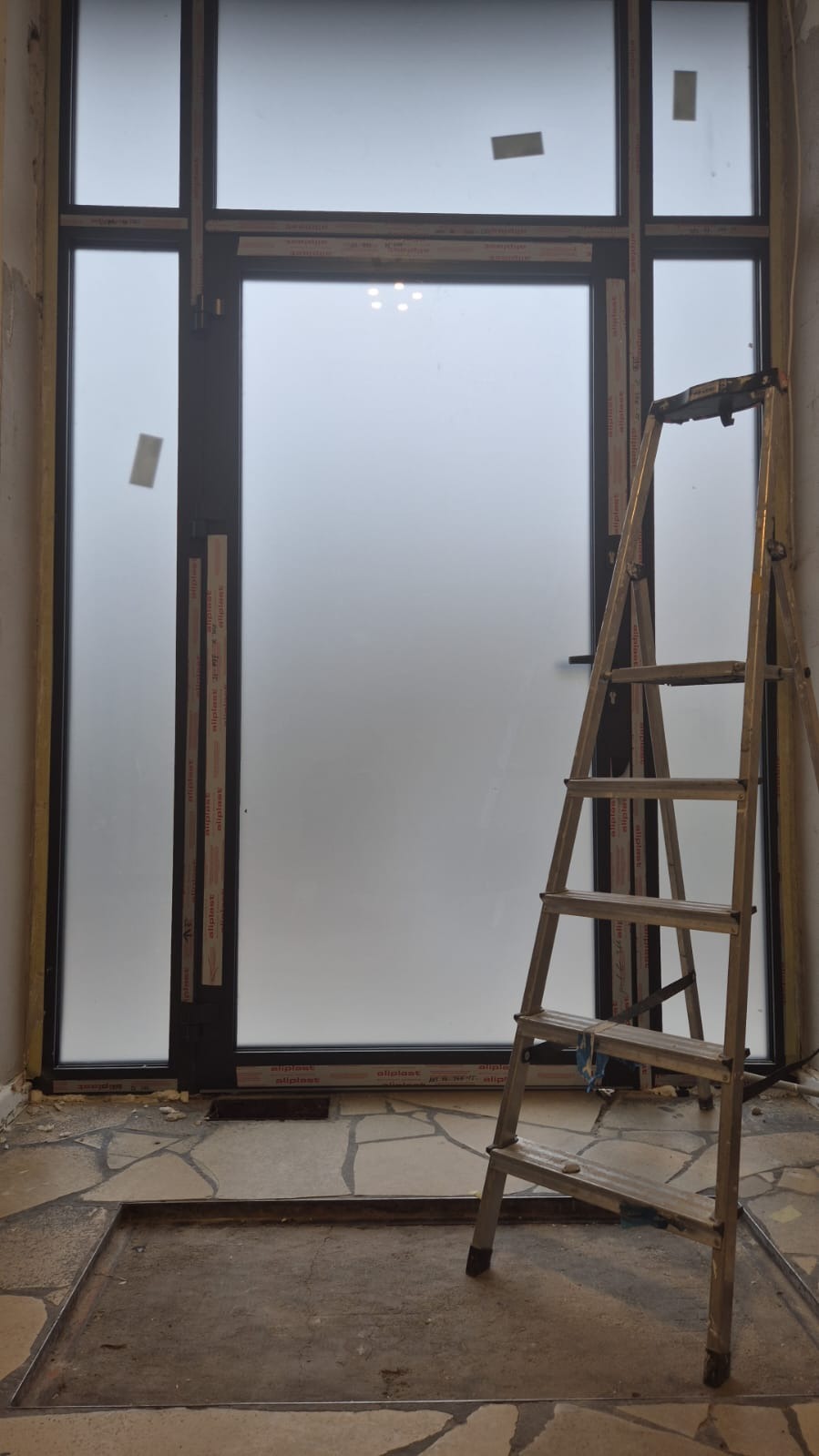 Montaż okna z matową szybą w surowym wnętrzu. Widoczna drabina, fragment podłogi z płytkami i ślady po pracach budowlanych. Okno z czarną ramą.