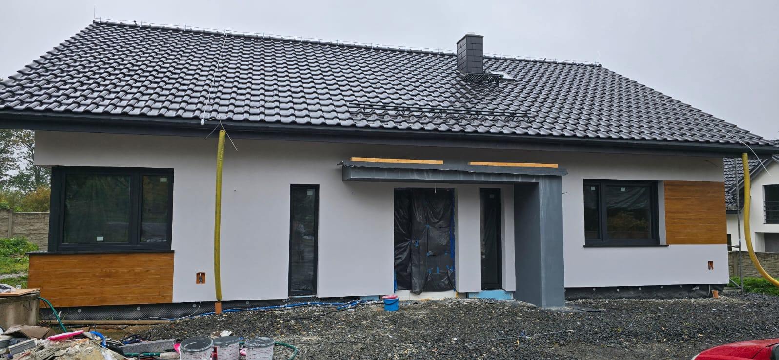 Nowoczesny dom jednorodzinny z ciemną dachówką i elementami dekoracyjnymi imitującymi drewno na elewacji. Widoczne detale wykończeniowe i teren wokół domu w trakcie prac.