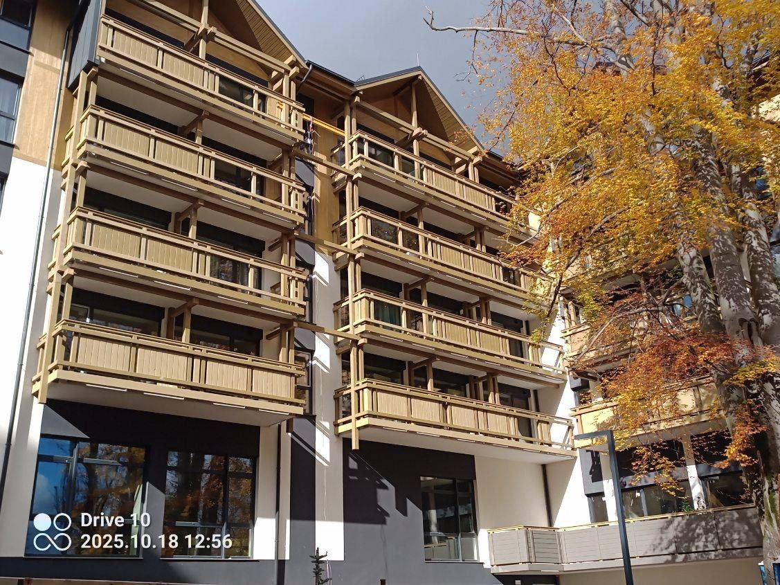 Wielopiętrowy budynek z drewnianymi balkonami w jesiennej scenerii. Uwagę przykuwa nowoczesna architektura i naturalne materiały wykończeniowe.