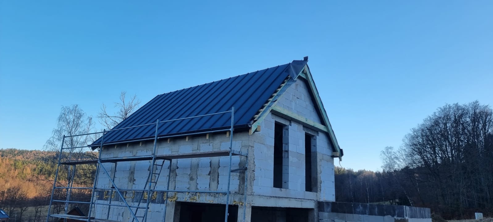 Niedokończony dom z ciemnym dachem z blachy na tle błękitnego nieba, widoczne rusztowania i drzewa w tle. Etap budowy domu z pustaków.
