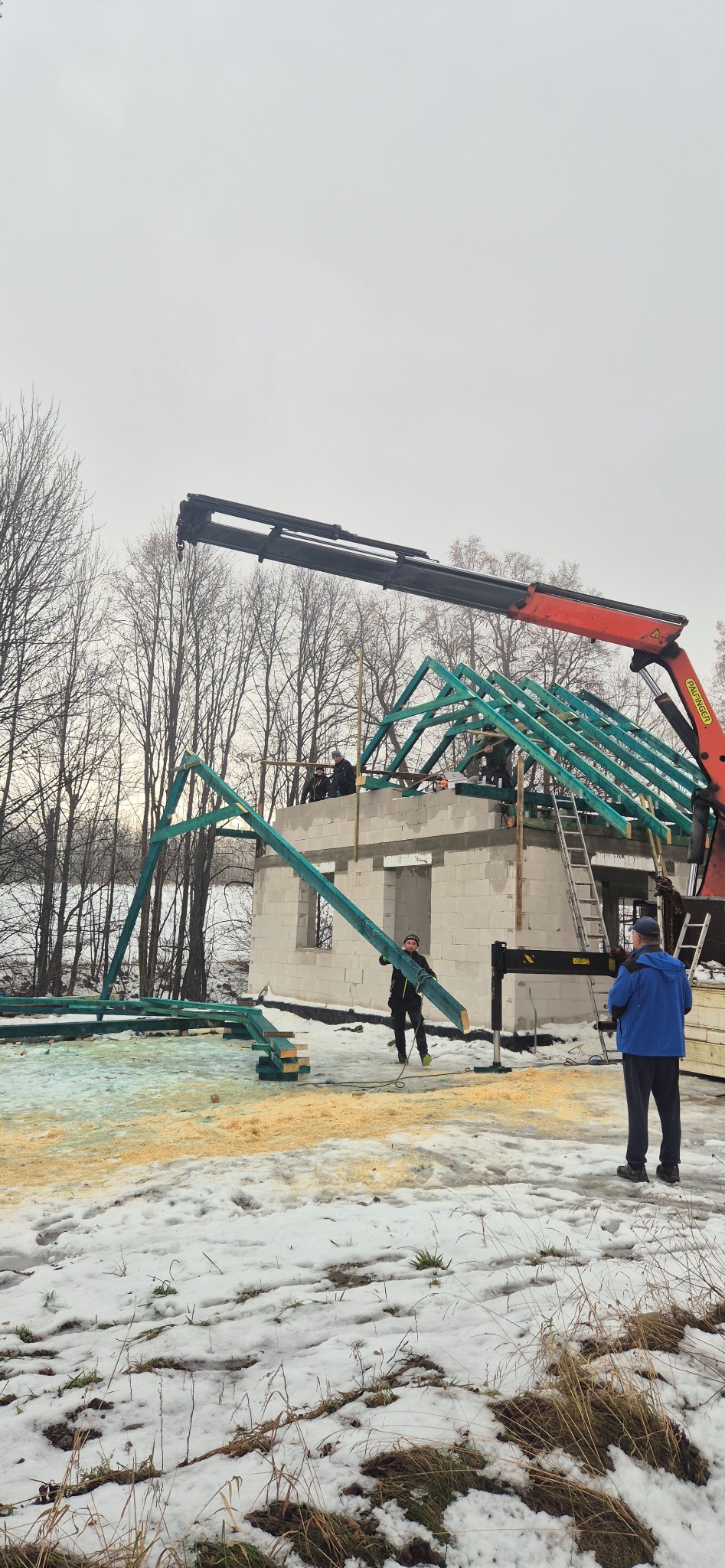 Budowa domu z bloczków: montaż zielonej więźby dachowej przy użyciu dźwigu. W tle drzewa i zimowy krajobraz. Widoczni pracownicy w trakcie pracy.