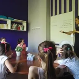 Sala lekcyjna z grupą dzieci siedzących przy drewnianym stole. Nauczycielka stoi przy białej tablicy, na której widać zapisane słowa. W tle fioletowa ściana z białymi pionowymi pasami.