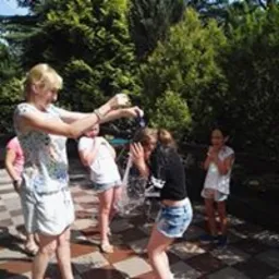 Grupa dzieci i dorosłych podczas letniej zabawy na świeżym powietrzu. Dorosła osoba w białej koszulce i krótkich spodenkach polewa wodą z wiadra młodą dziewczynę w dżinsowych szortach i czarnej...