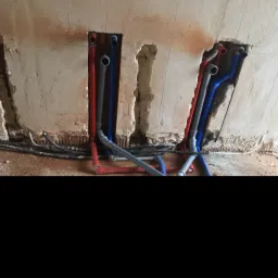 Wykute w ścianie bruzdy z instalacją wodną i rurami grzewczymi PEX w kolorach czerwonym i niebieskim, oraz szarymi rurami kanalizacyjnymi. Widoczne przewody elektryczne w peszlach. Ściana w stanie...