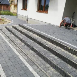 Układanie ciemnoszarej kostki brukowej na schodach wejściowych do domu, z jasnymi fugami i obrzeżami, pracownik wygładza powierzchnię.