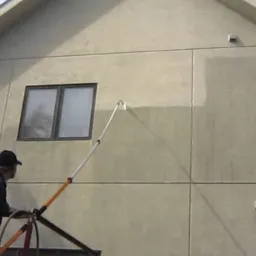 Pracownik na rusztowaniu czyści elewację budynku za pomocą lancy teleskopowej; widoczna wyraźna różnica w kolorze pomiędzy umytą a brudną powierzchnią.