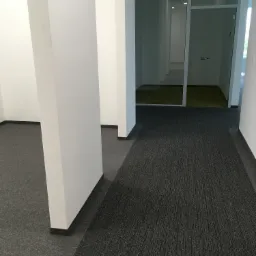 Szara wykładzina dywanowa w biurze z białymi filarami i przeszklonymi pomieszczeniami w tle.