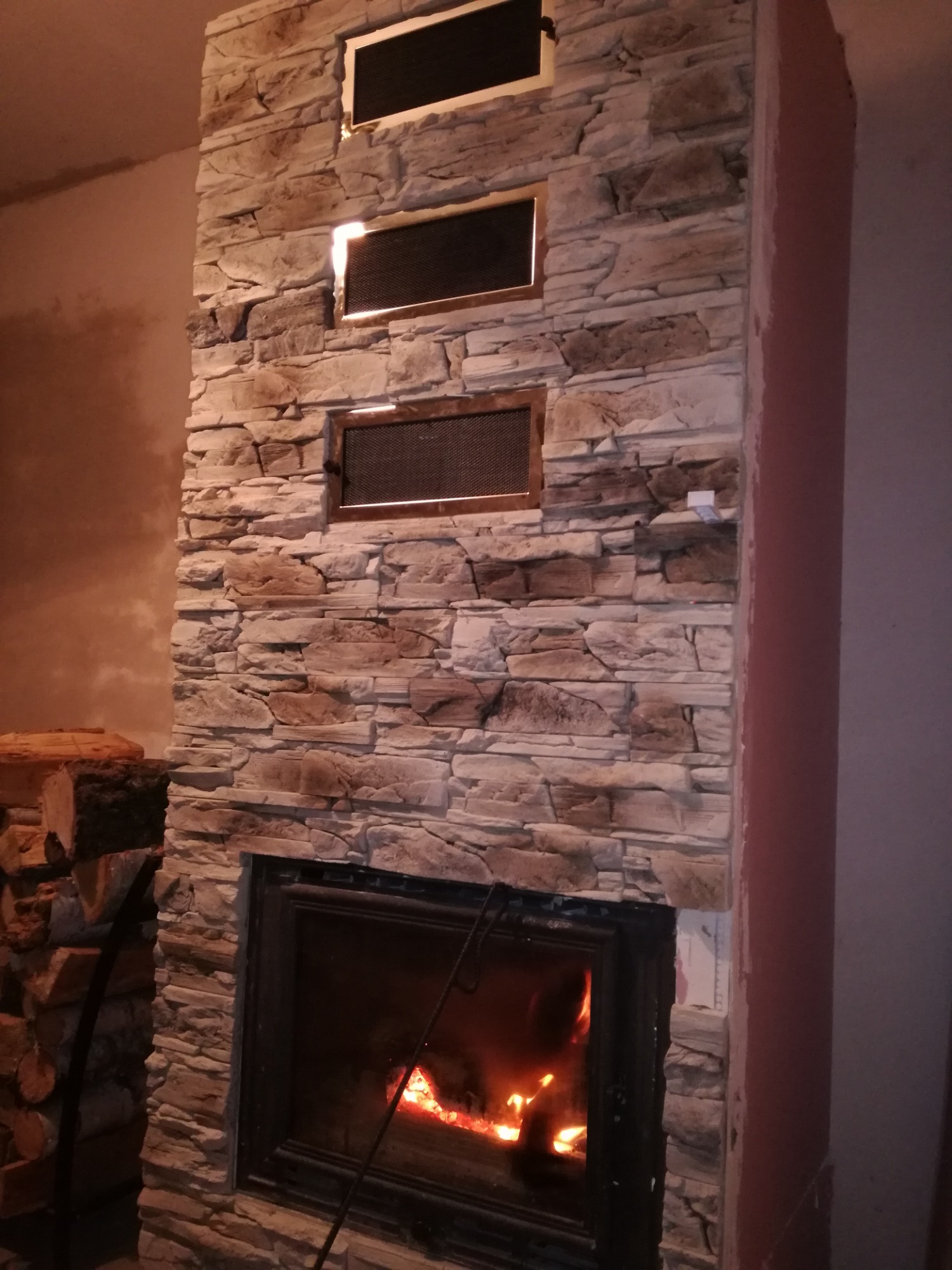 Kominek obłożony kamieniem dekoracyjnym z trzema kratkami wentylacyjnymi powyżej paleniska, obok ułożone drewno do kominka.