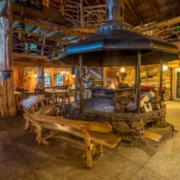 Bukowa Chata - Wnętrze przestronnej restauracji w stylu góralskim z centralnie umieszczonym paleniskiem otoczonym kamienną konstrukcją, drewnianymi ławami i stołami w tle.