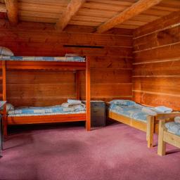 Bukowa Chata - Rustykalny pokój z drewnianymi ścianami, łóżkiem piętrowym i dwoma pojedynczymi łóżkami z niebieskimi narzutami, stolikiem i krzesłami oraz łapaczem snów na ścianie.