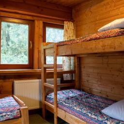 Bukowa Chata - Wnętrze drewnianego domku z dwoma łóżkami, jedno nad drugim, z wzorzystą pościelą, widok na okno z zasłonką w kwiaty i lampką nocną na parapecie.