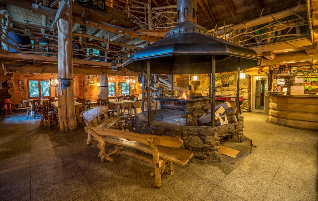 Wnętrze przestronnej restauracji w stylu góralskim z centralnie umieszczonym paleniskiem otoczonym kamienną konstrukcją, drewnianymi ławami i stołami w tle.