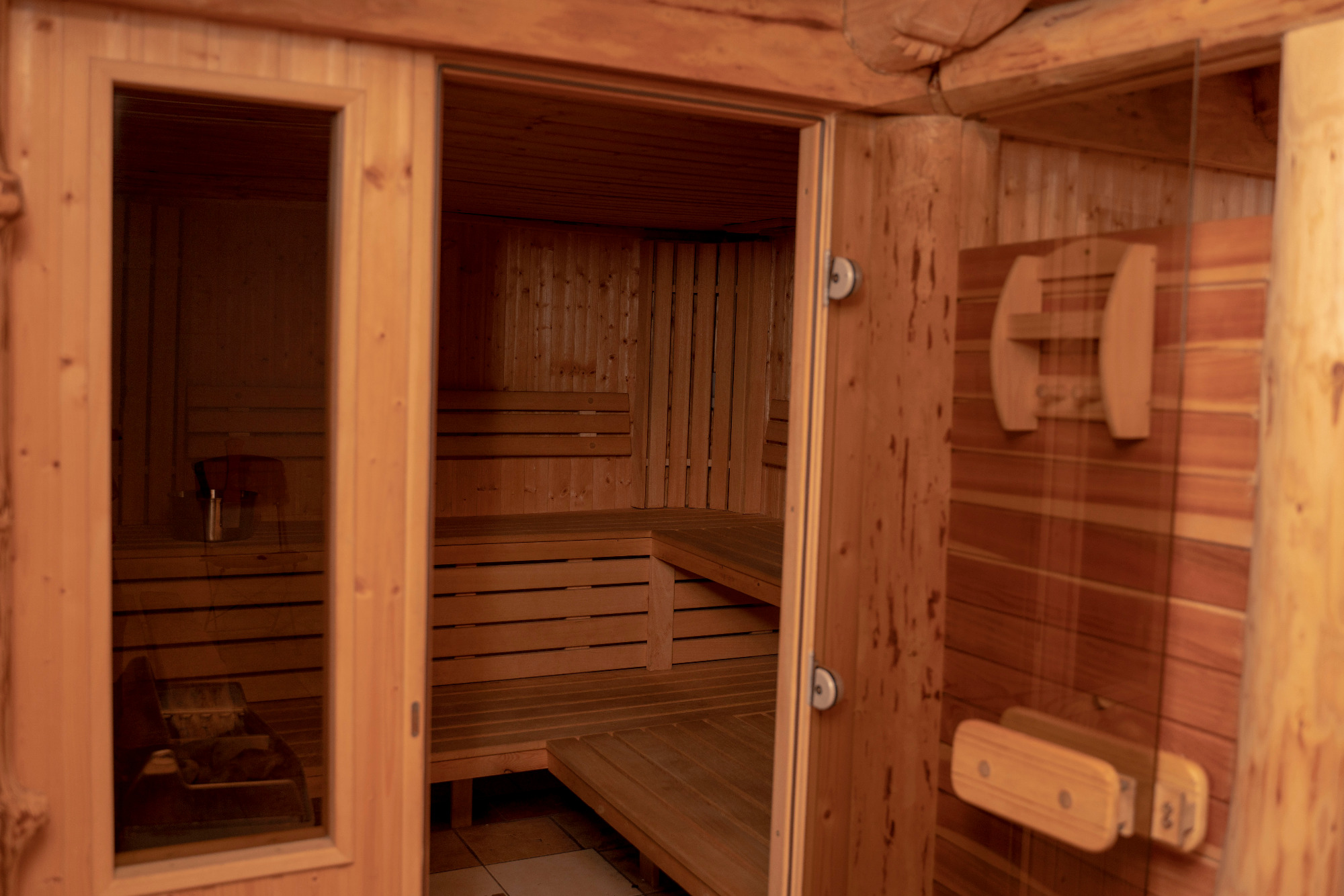 Wnętrze drewnianej sauny z ławkami i szklanymi drzwiami, widok z zewnątrz przez otwarte drzwi.