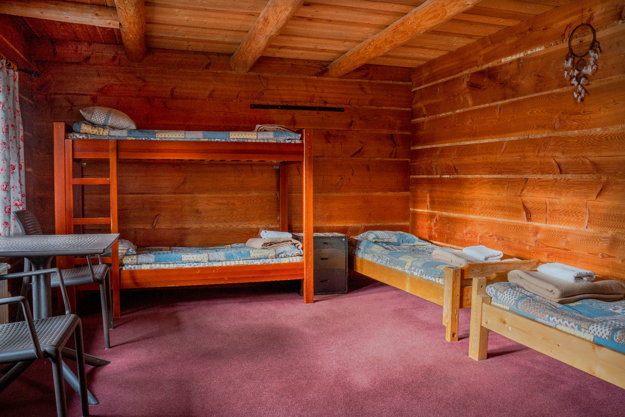 Rustykalny pokój z drewnianymi ścianami, łóżkiem piętrowym i dwoma pojedynczymi łóżkami z niebieskimi narzutami, stolikiem i krzesłami oraz łapaczem snów na ścianie.