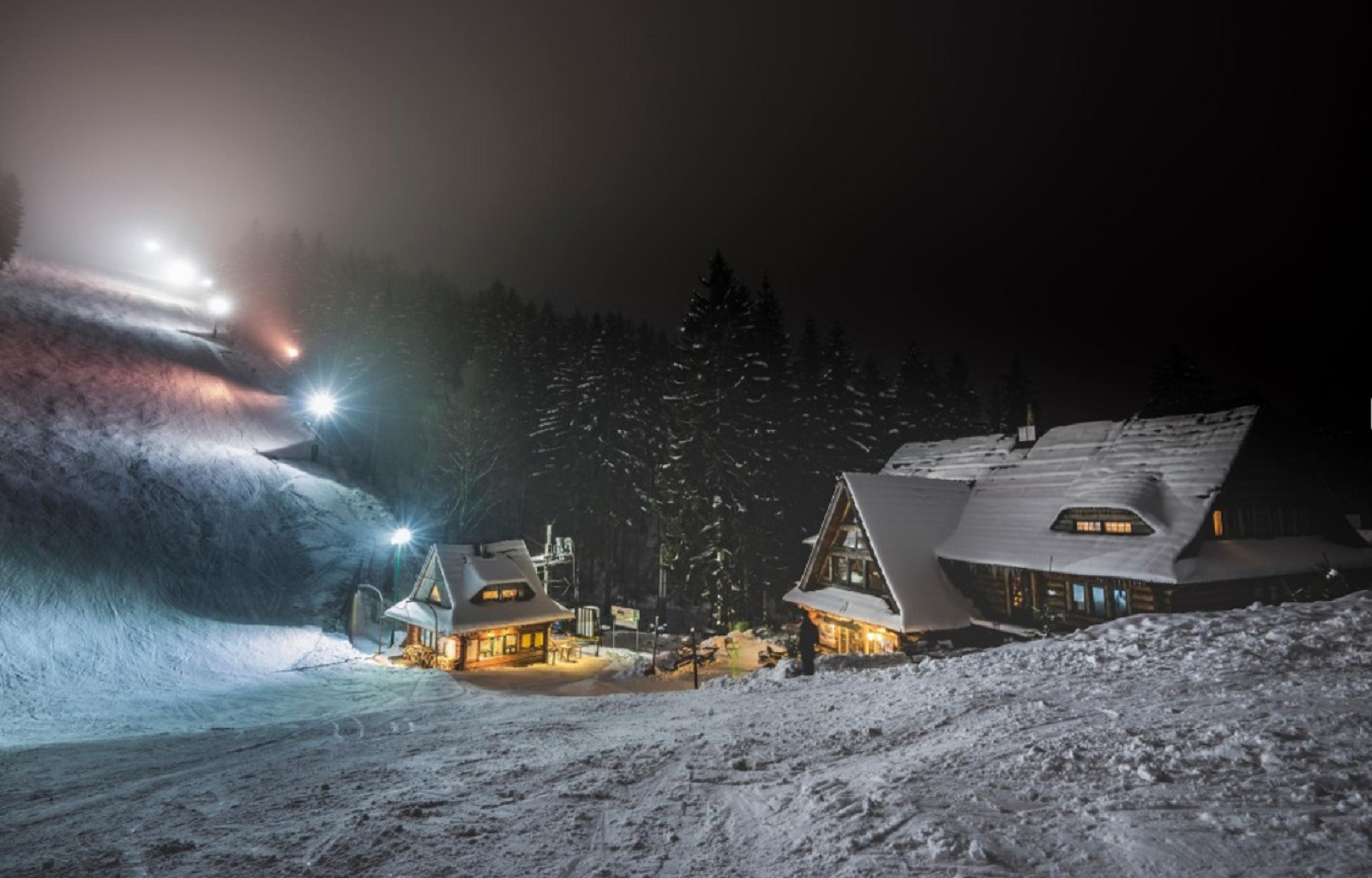 Ośnieżony stok narciarski nocą z oświetlonymi budynkami w stylu góralskim i lasem w tle.