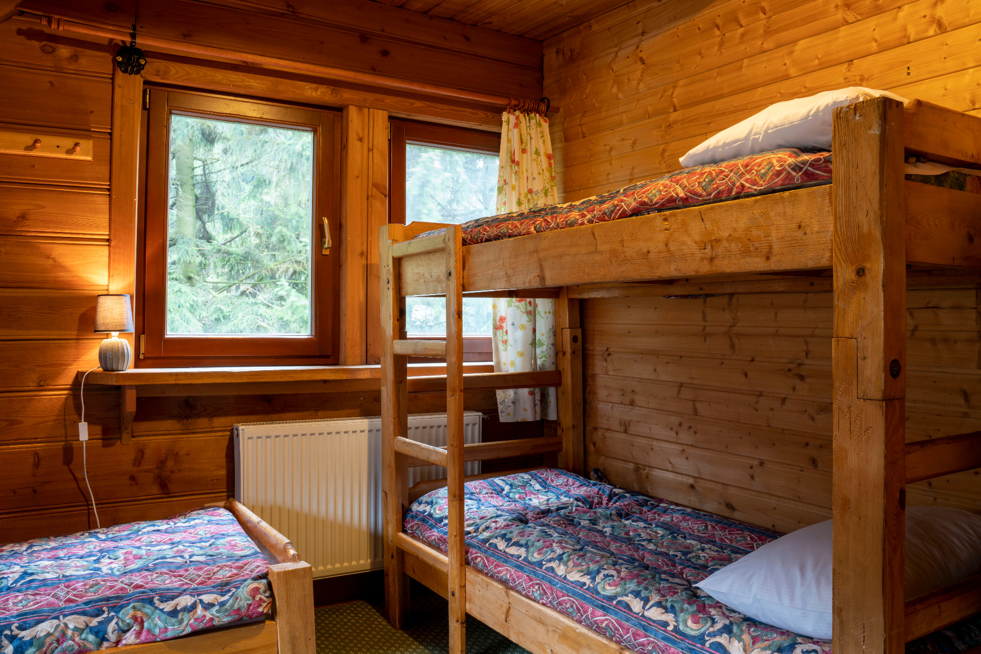 Wnętrze drewnianego domku z dwoma łóżkami, jedno nad drugim, z wzorzystą pościelą, widok na okno z zasłonką w kwiaty i lampką nocną na parapecie.