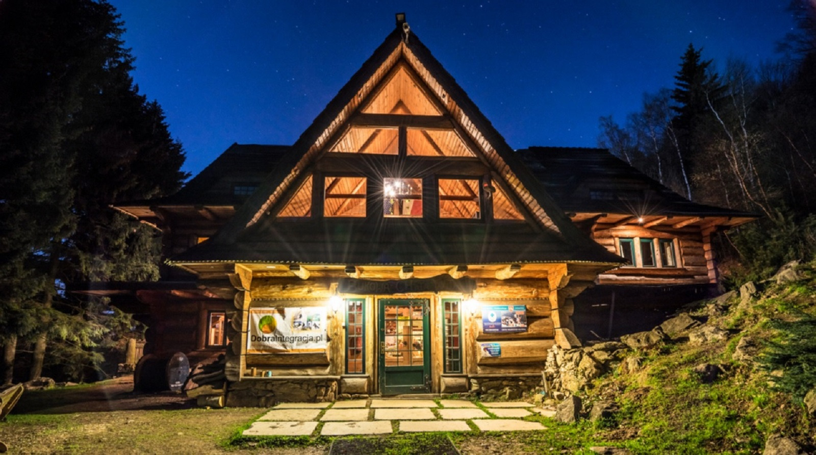 Oświetlony, drewniany budynek w stylu góralskim z trójkątnym dachem i jasnymi oknami, widoczny nocą na tle ciemnego nieba z gwiazdami, otoczony drzewami i zielenią.