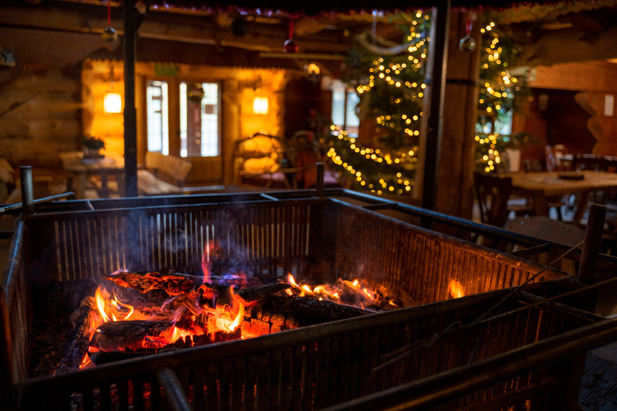 Ognisko w metalowej skrzyni w rustykalnym wnętrzu z choinką w tle, płomienie oświetlają otoczenie