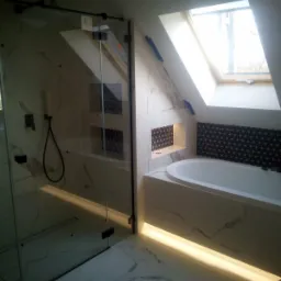 Nowoczesna łazienka na poddaszu z kabiną prysznicową typu walk-in, wykończona białym marmurem i czarną mozaiką, oświetlona taśmami LED, z oknem dachowym w skosie.