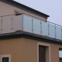 Balkon z nowoczesną, szklaną balustradą ze stalowymi słupkami, zamontowaną na beżowej elewacji budynku.