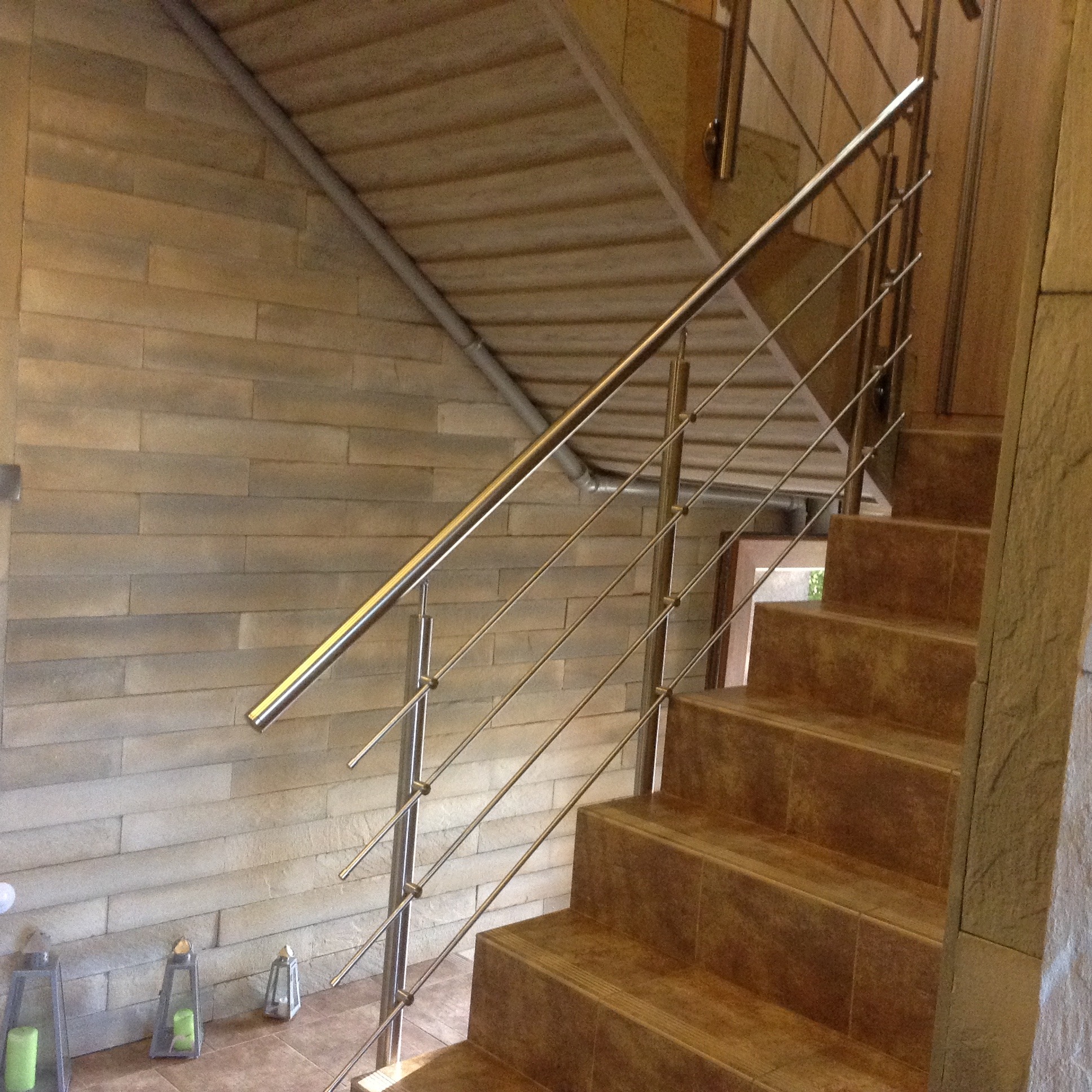 Schody wyłożone brązowymi płytkami z nierdzewną, minimalistyczną barierką z poziomych prętów. W tle ściana z kamiennych płytek i dekoracyjne lampiony pod schodami.