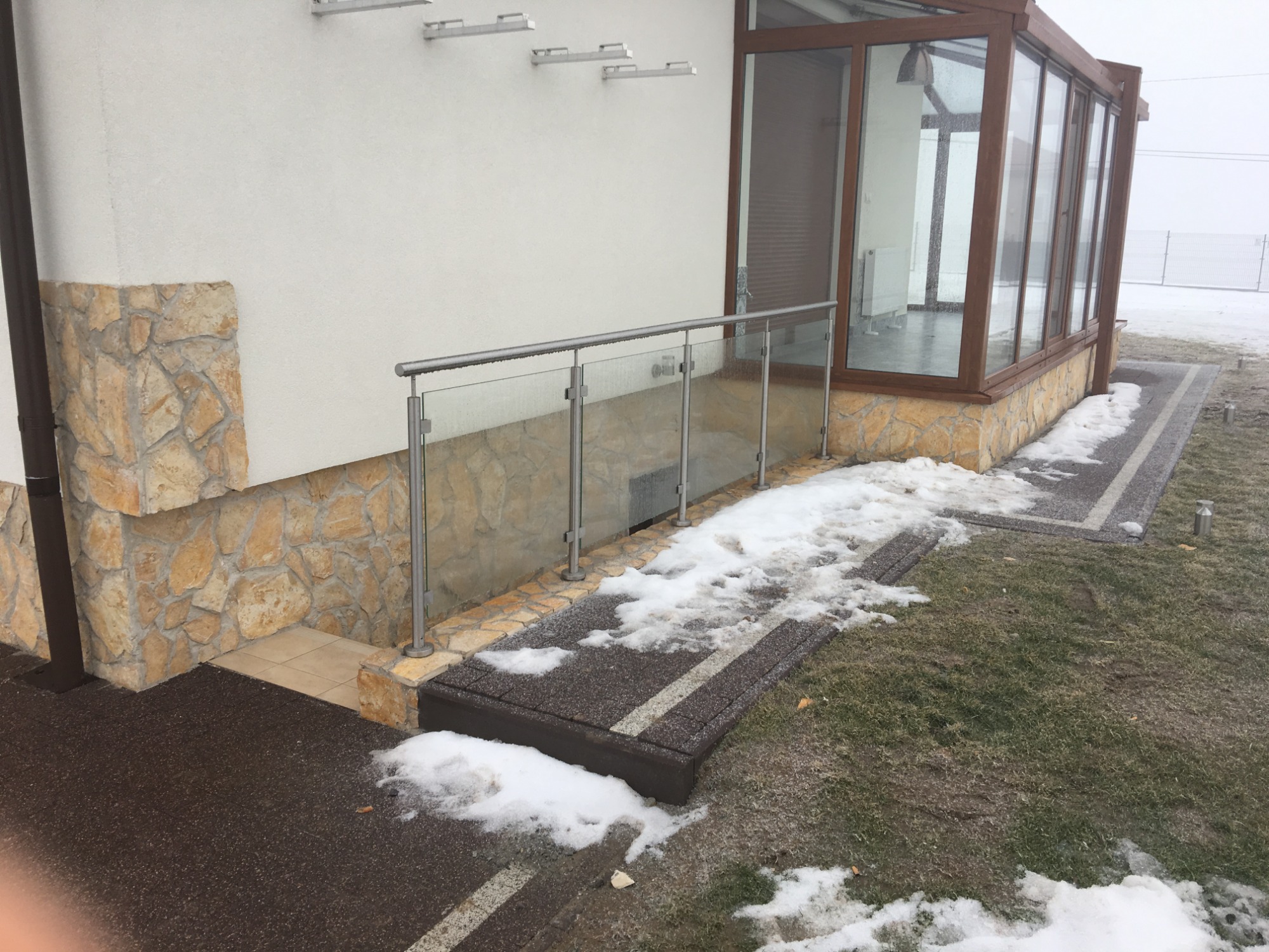 Szklana barierka z metalowymi słupkami zabezpieczająca zejście do piwnicy przy budynku z kamienną elewacją, częściowo przykryta śniegiem.