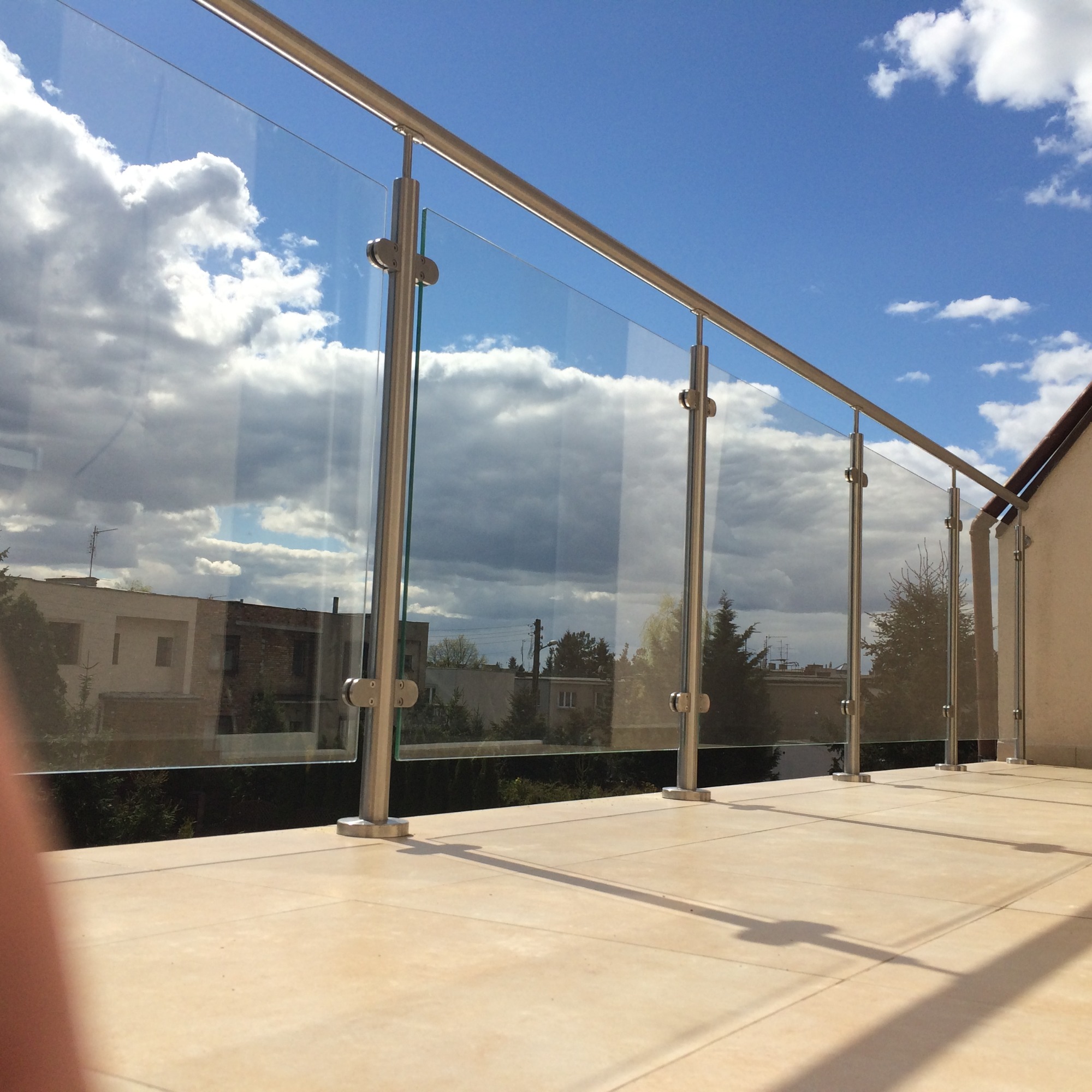 Szklana balustrada balkonowa ze stali nierdzewnej, mocowana punktowo, z widokiem na budynki i zachmurzone niebo.