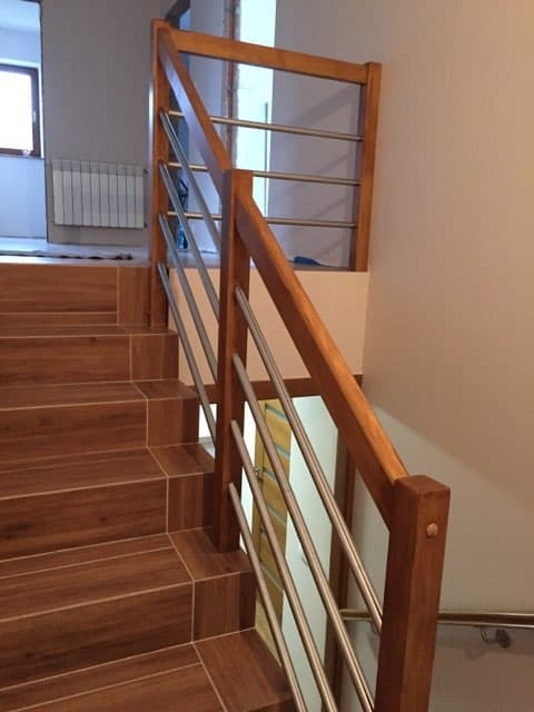 Schody wyłożone brązowymi płytkami imitującymi drewno, zabezpieczone balustradą z drewnianymi słupkami i poziomymi, metalowymi prętami.