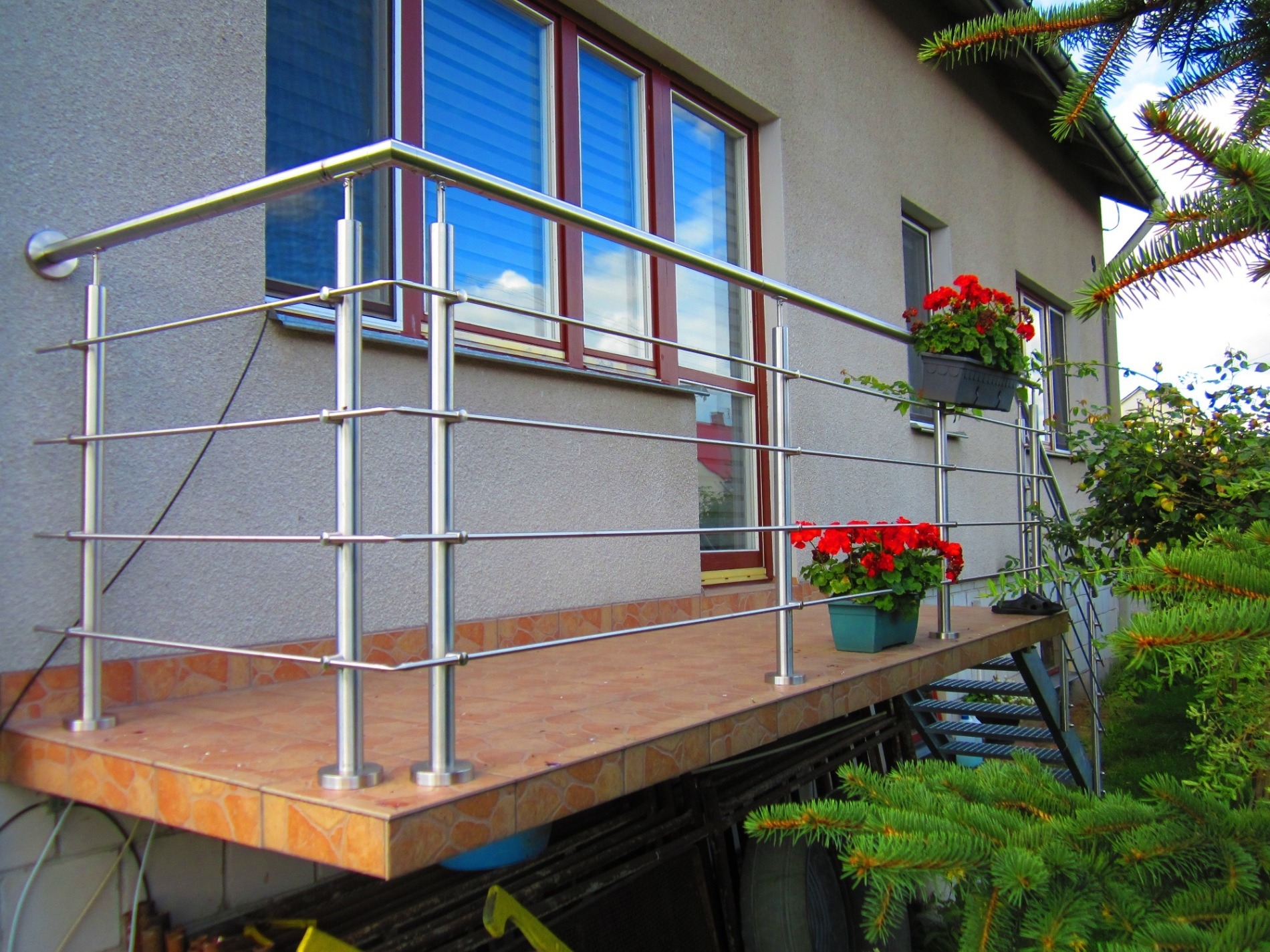 Balkon z chromowaną, minimalistyczną balustradą z poziomych prętów, wyłożony terakotą, z czerwonymi kwiatami w donicach, widok z dołu na tle ściany budynku i igliwia.