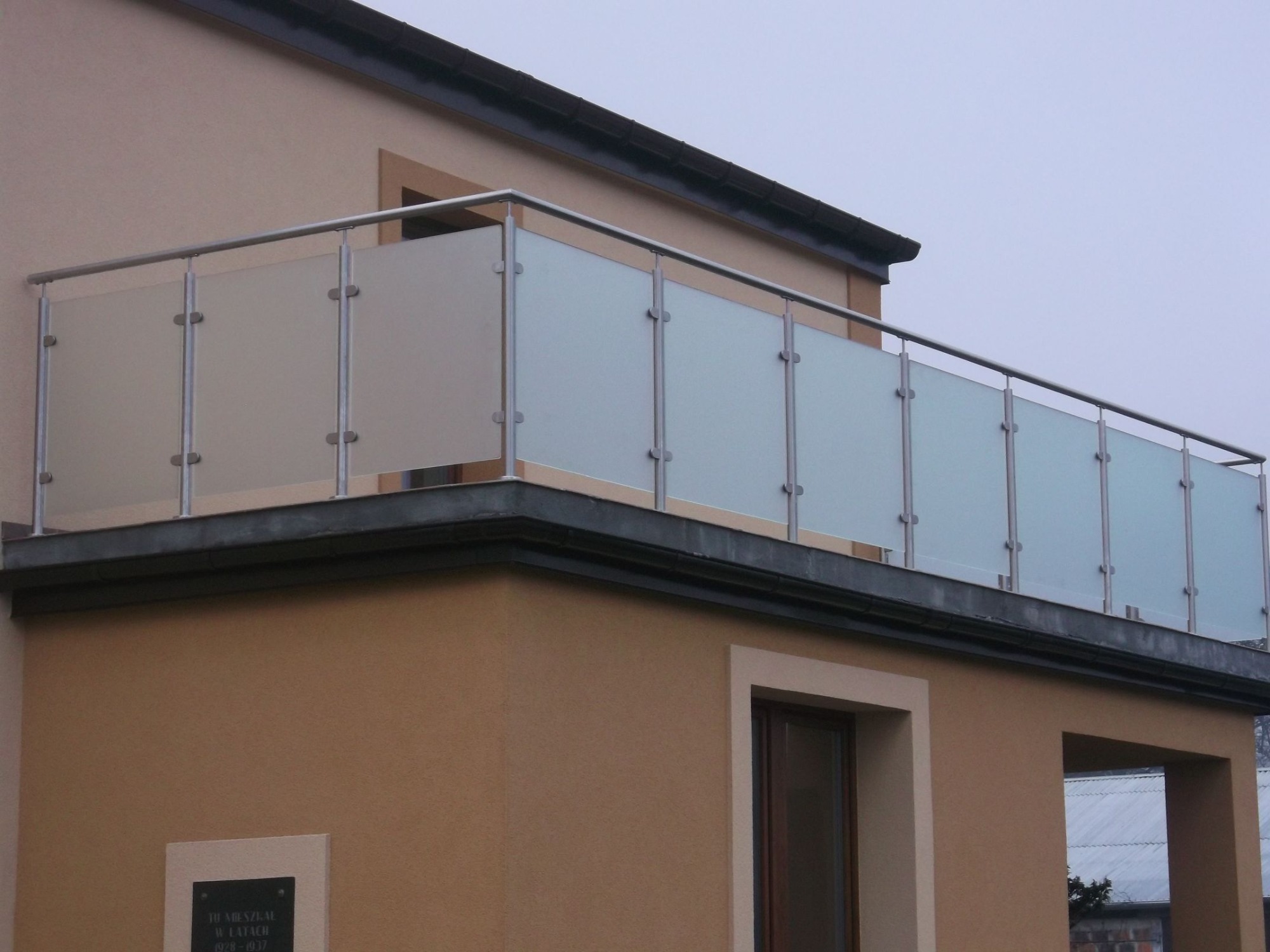 Balkon z nowoczesną, szklaną balustradą ze stalowymi słupkami, zamontowaną na beżowej elewacji budynku.