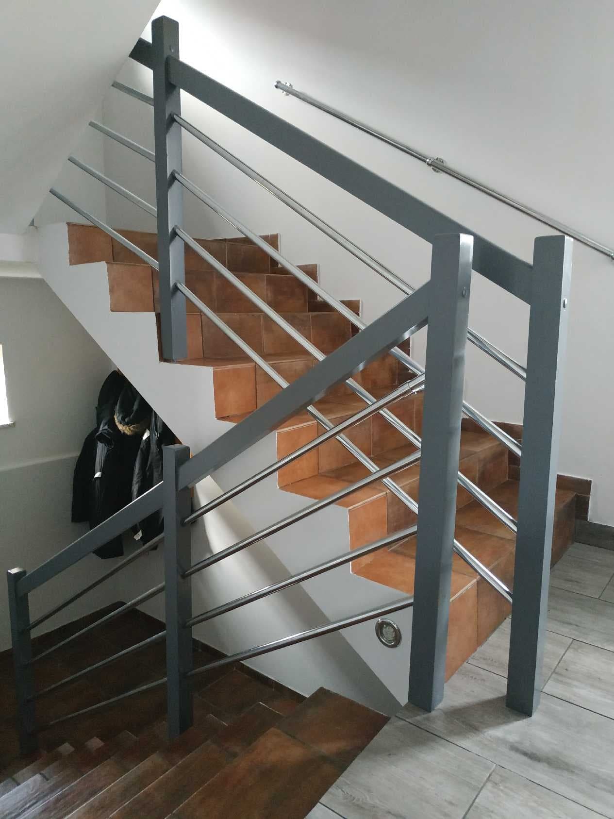 Schody z terakoty z szarą, metalową konstrukcją zabezpieczającą i metalową poręczą na ścianie, widok z dołu.