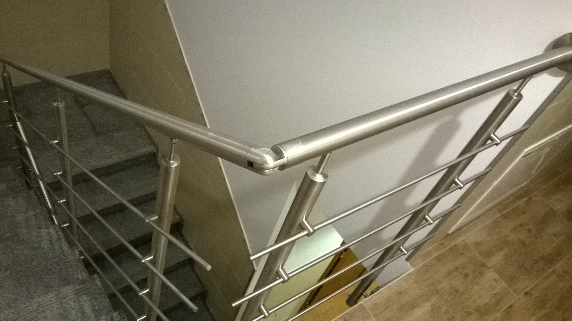Szczegółowe ujęcie nierdzewnej poręczy schodowej z poziomymi prętami, mocowanej do słupków i ściany, widok z góry na schody wyłożone szarymi płytkami.